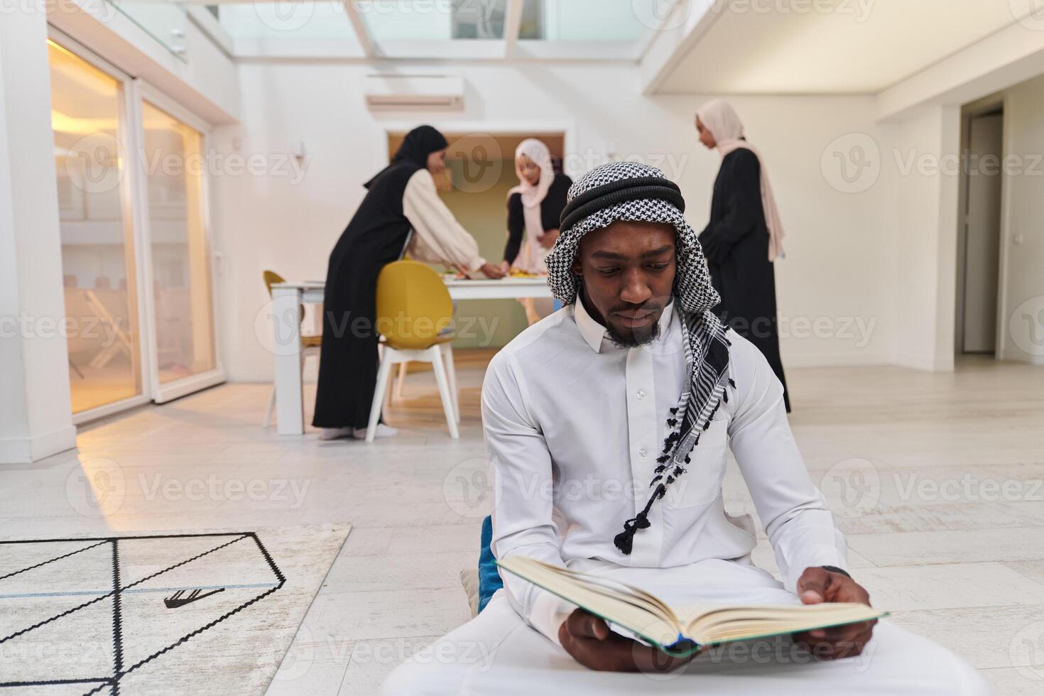 In the sacred month of Ramadan, an African American Muslim man engrossed in reading the Holy Quran is surrounded by a backdrop where three hijab-wearing women prepare and serve food for iftar photo