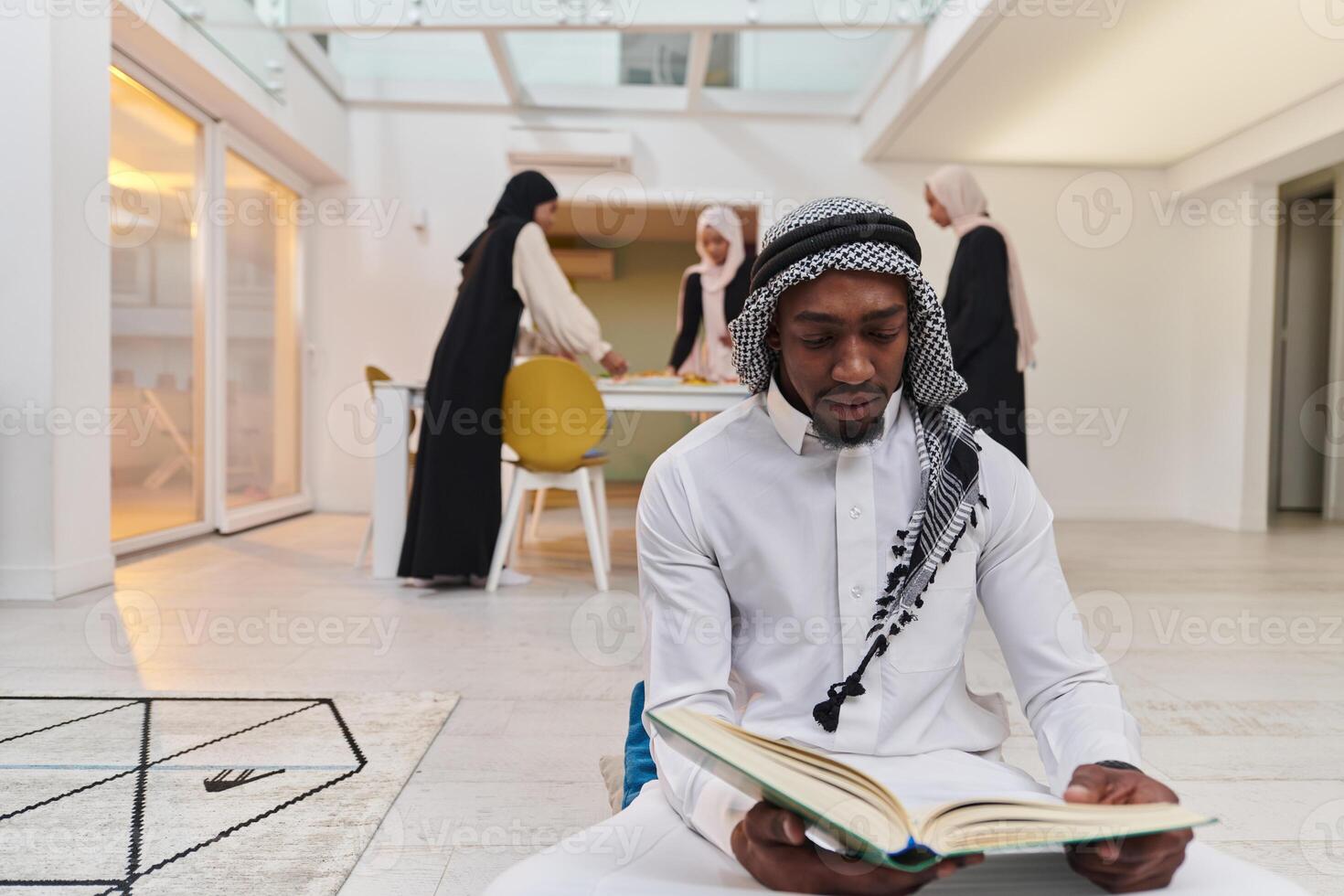 In the sacred month of Ramadan, an African American Muslim man engrossed in reading the Holy Quran is surrounded by a backdrop where three hijab-wearing women prepare and serve food for iftar photo