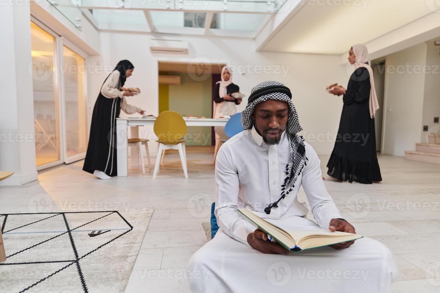 In the sacred month of Ramadan, an African American Muslim man engrossed in reading the Holy Quran is surrounded by a backdrop where three hijab-wearing women prepare and serve food for iftar photo