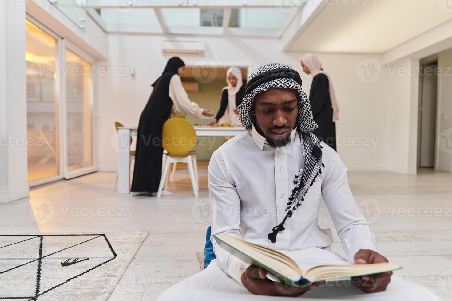 In the sacred month of Ramadan, an African American Muslim man engrossed in reading the Holy Quran is surrounded by a backdrop where three hijab-wearing women prepare and serve food for iftar photo