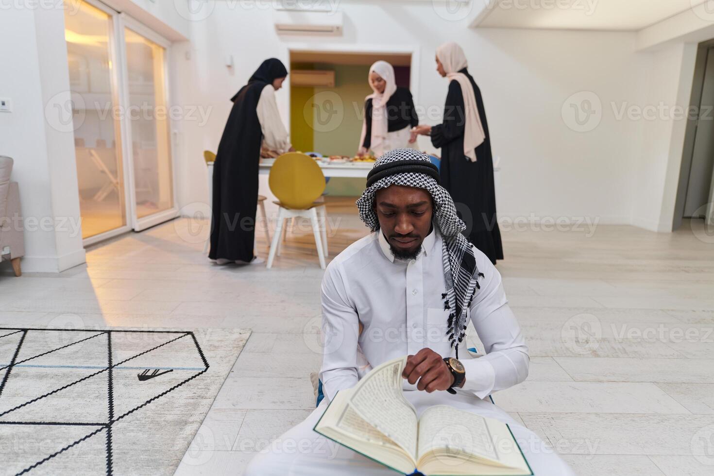 In the sacred month of Ramadan, an African American Muslim man engrossed in reading the Holy Quran is surrounded by a backdrop where three hijab-wearing women prepare and serve food for iftar photo