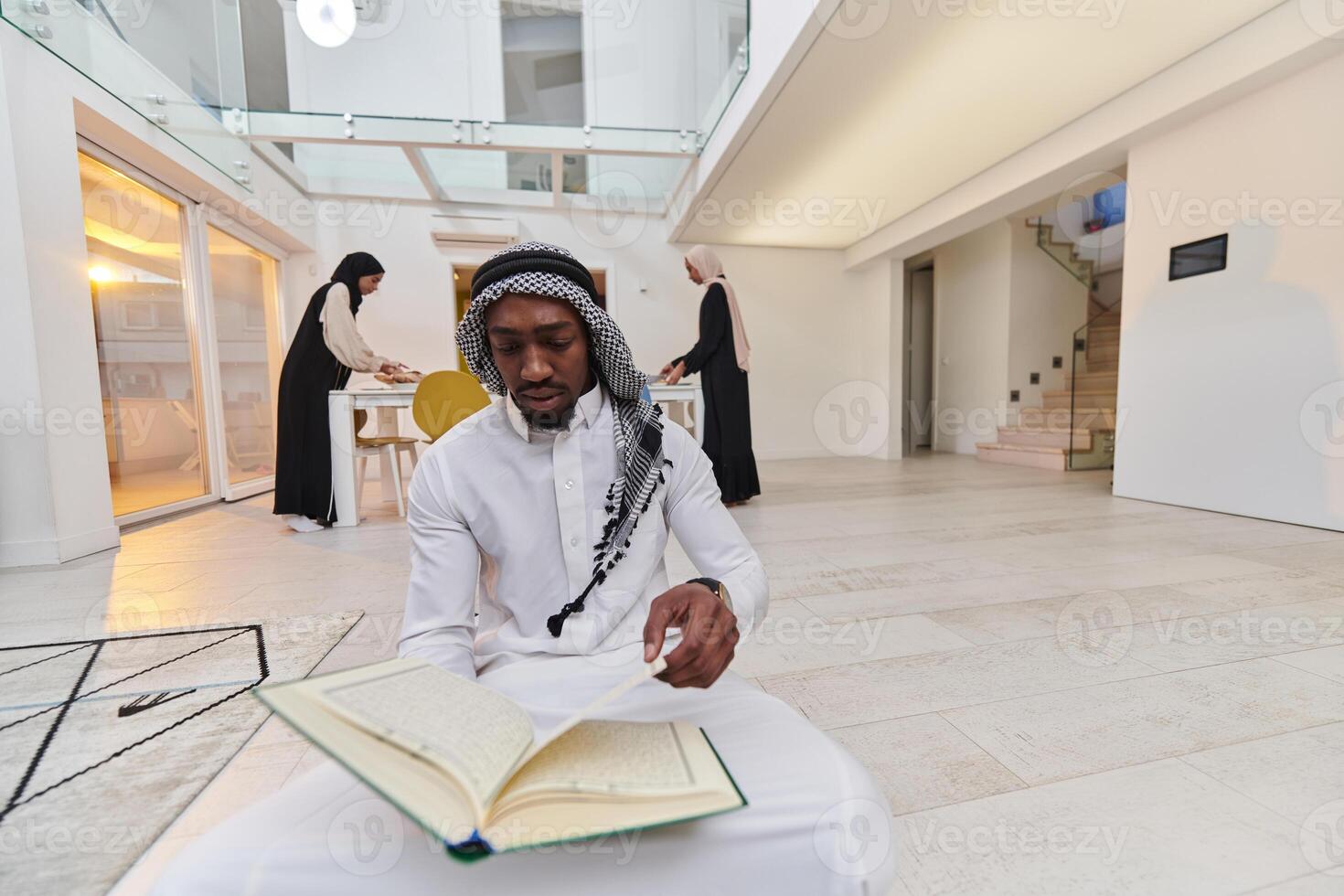 In the sacred month of Ramadan, an African American Muslim man engrossed in reading the Holy Quran is surrounded by a backdrop where three hijab-wearing women prepare and serve food for iftar photo