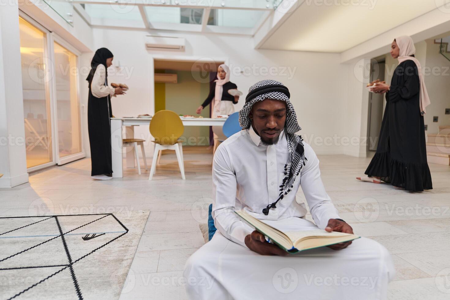 In the sacred month of Ramadan, an African American Muslim man engrossed in reading the Holy Quran is surrounded by a backdrop where three hijab-wearing women prepare and serve food for iftar photo