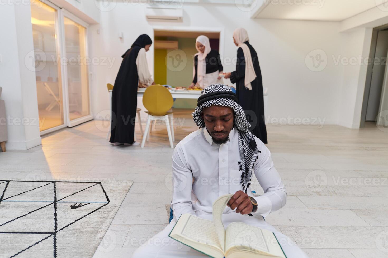 In the sacred month of Ramadan, an African American Muslim man engrossed in reading the Holy Quran is surrounded by a backdrop where three hijab-wearing women prepare and serve food for iftar photo