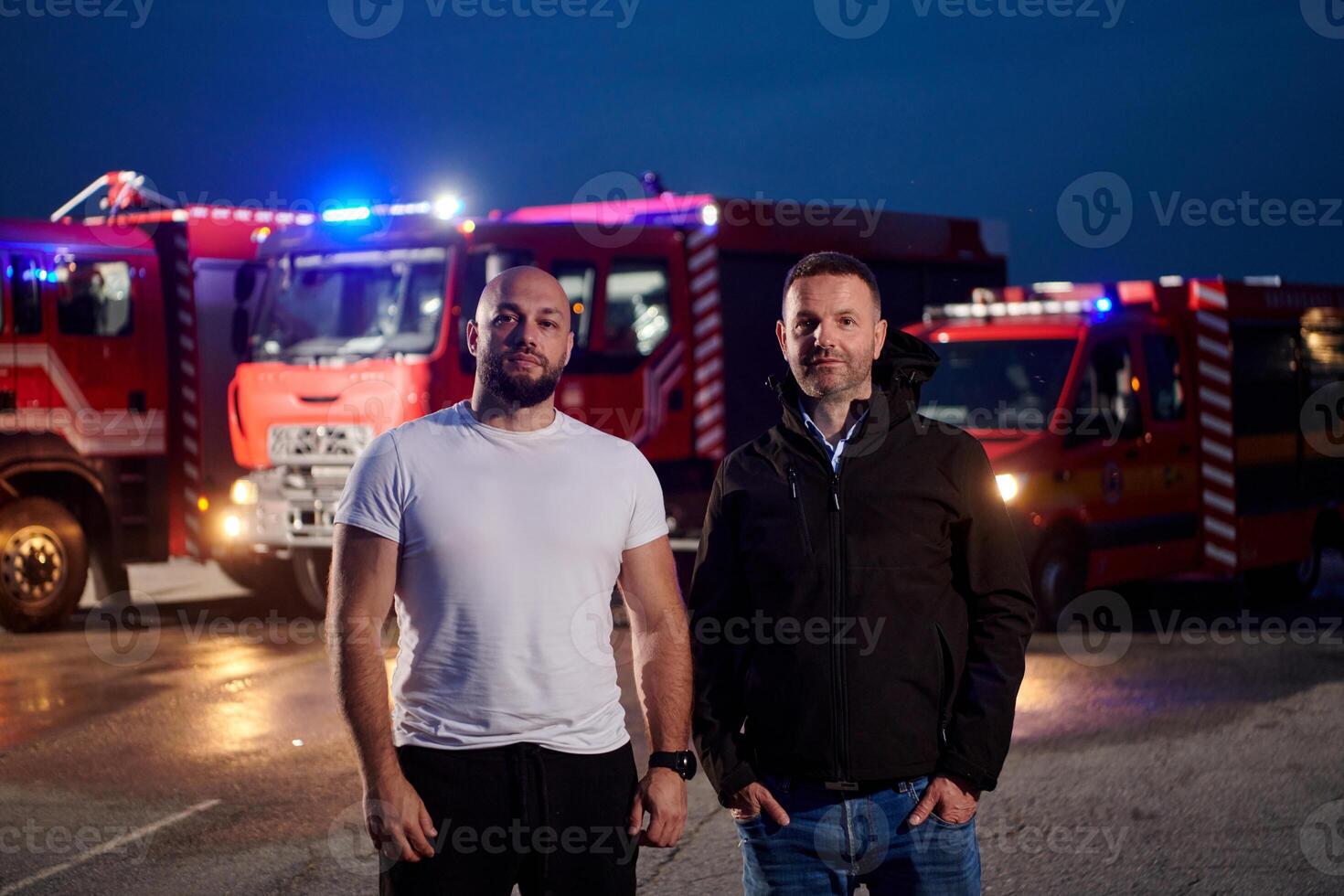 Group of firefighters, dressed in civilian clothing, stand in front of fire trucks during the night, showcasing a moment of camaraderie and unity among the team as they reflect on their duties and the photo