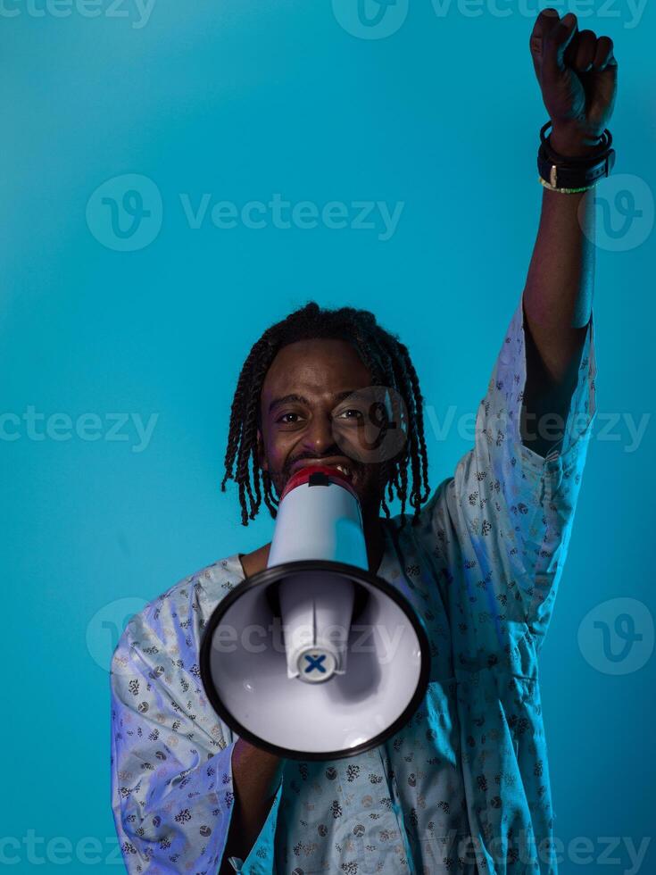 In a powerful and symbolic image, an African American man wears traditional clothing, passionately wields a megaphone against a striking blue background, with his hand raised in the air symbolizing photo
