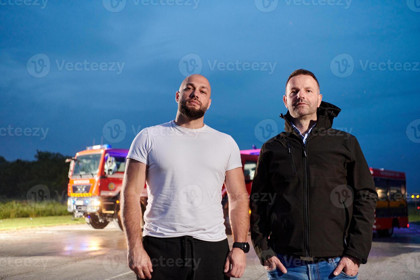 Group of firefighters, dressed in civilian clothing, stand in front of fire trucks during the night, showcasing a moment of camaraderie and unity among the team as they reflect on their duties and the photo