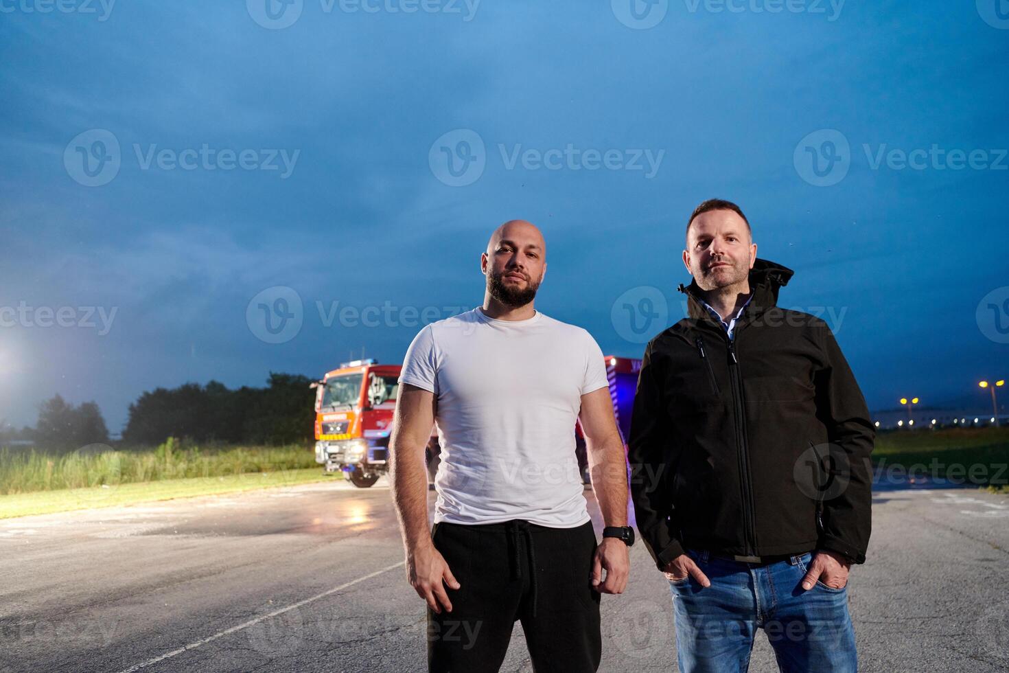 Group of firefighters, dressed in civilian clothing, stand in front of fire trucks during the night, showcasing a moment of camaraderie and unity among the team as they reflect on their duties and the photo