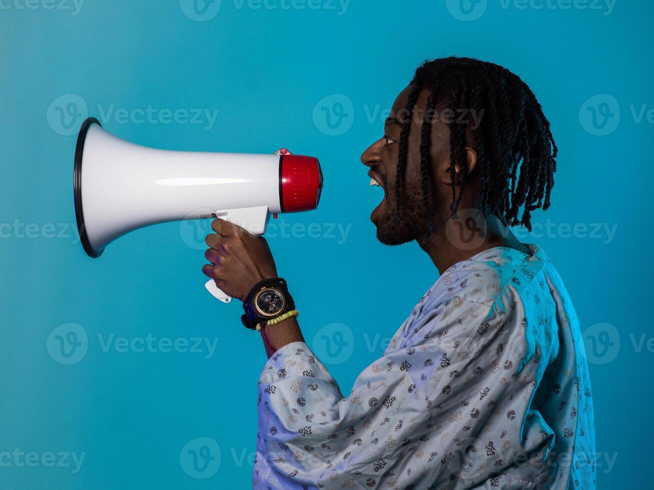 African American man dons traditional attire, passionately utilizing a megaphone against a striking blue background, symbolizing his vocal and cultural empowerment in the pursuit of social justice and photo