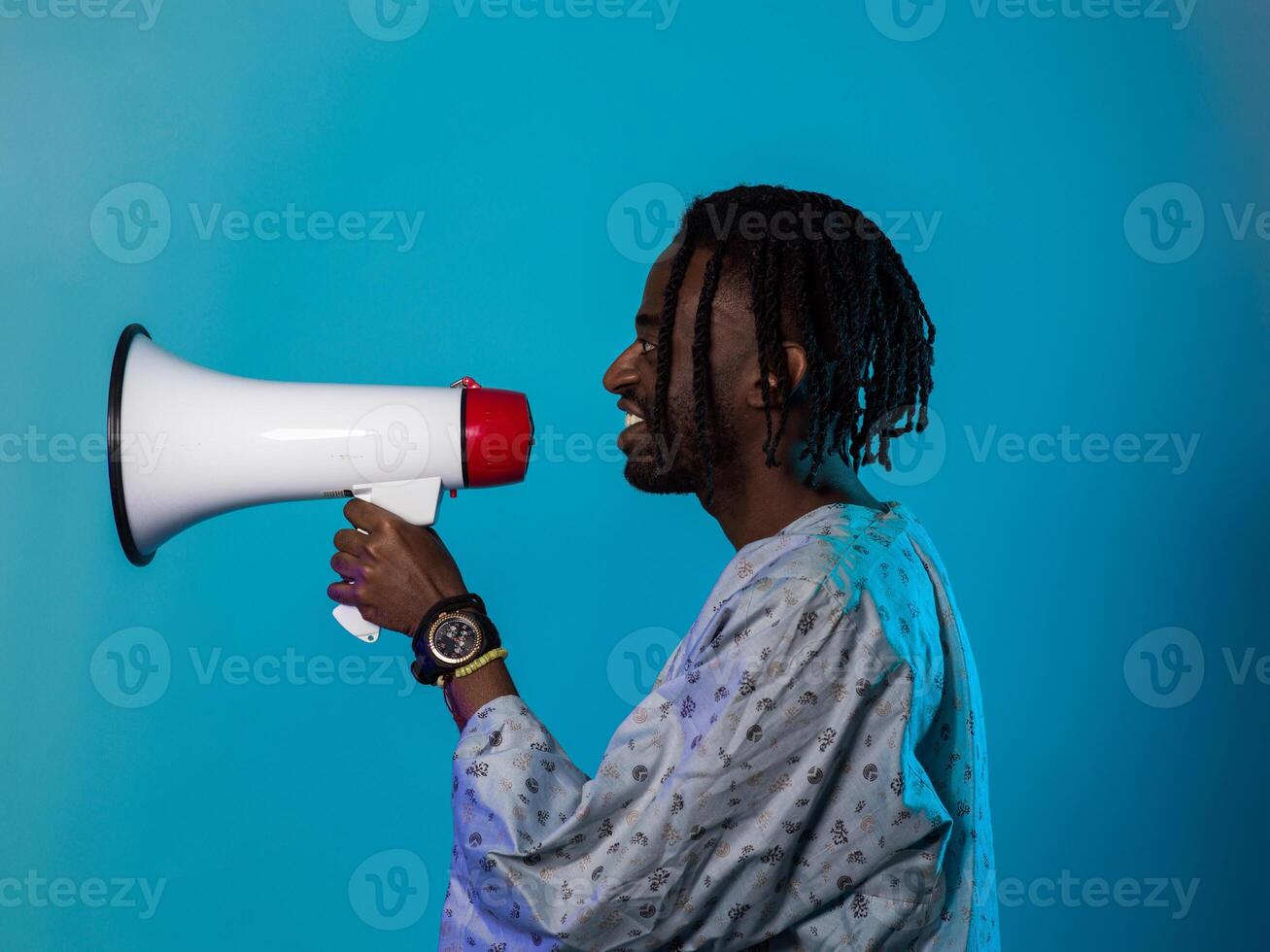African American man dons traditional attire, passionately utilizing a megaphone against a striking blue background, symbolizing his vocal and cultural empowerment in the pursuit of social justice and photo