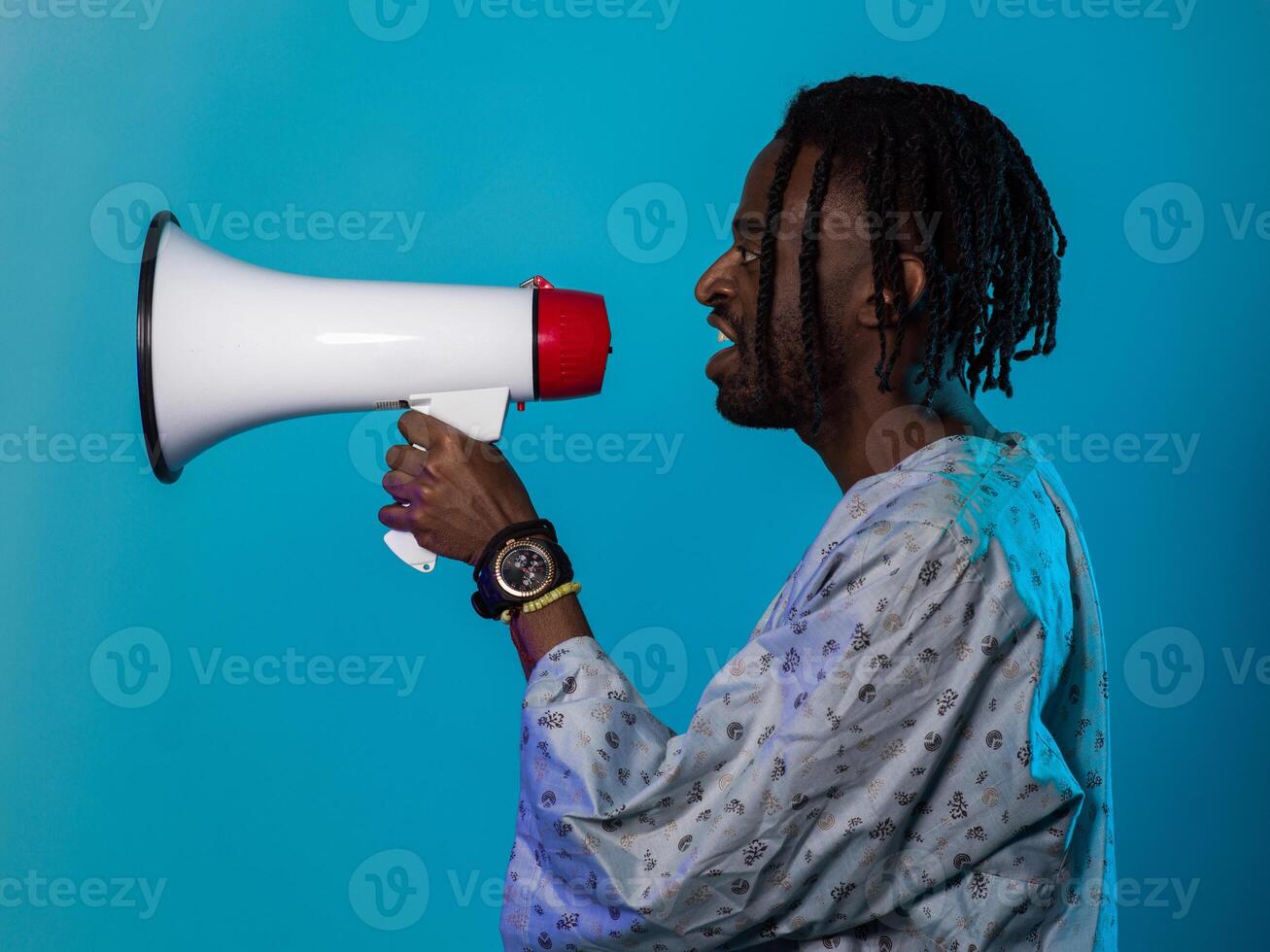 African American man dons traditional attire, passionately utilizing a megaphone against a striking blue background, symbolizing his vocal and cultural empowerment in the pursuit of social justice and photo
