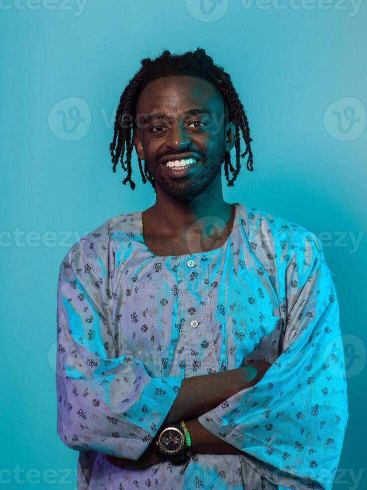 A Sudanese man adorned with modern dreadlocks stands proudly in traditional Sudanese attire, his arms crossed, conveying a blend of cultural heritage and contemporary style against a vibrant blue photo
