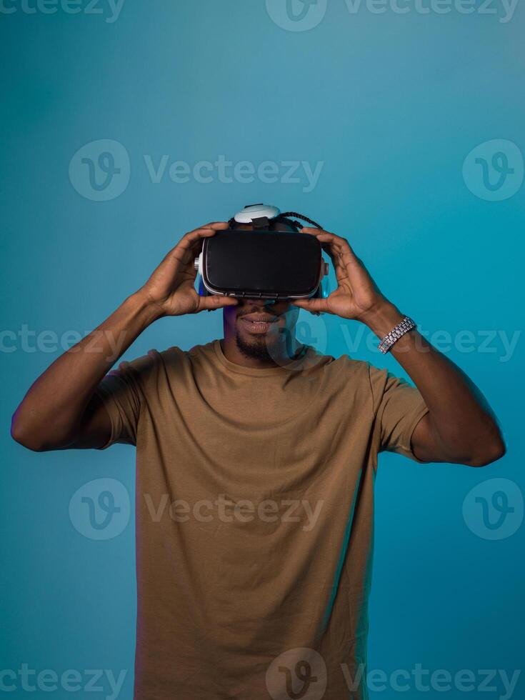 In a futuristic visual, an African American man stands isolated against a striking blue backdrop, adorned with VR glasses that transport him into a cutting-edge virtual reality experience, merging photo