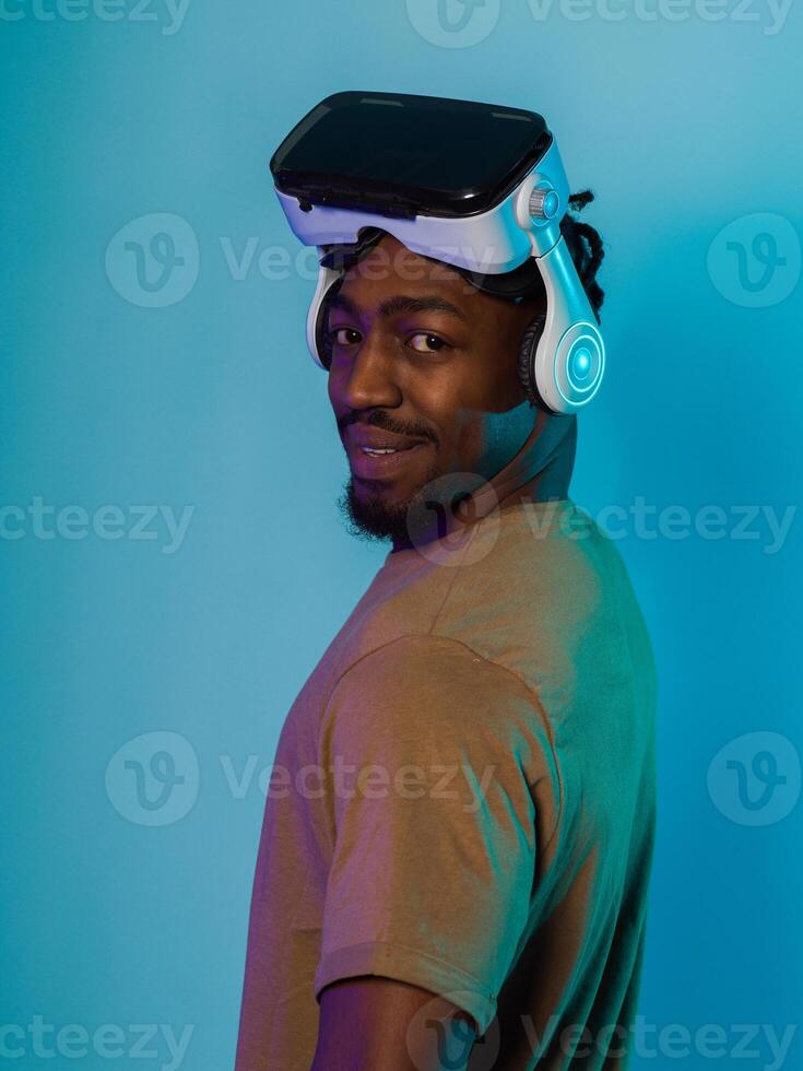 In a futuristic visual, an African American man stands isolated against a striking blue backdrop, adorned with VR glasses that transport him into a cutting-edge virtual reality experience, merging photo
