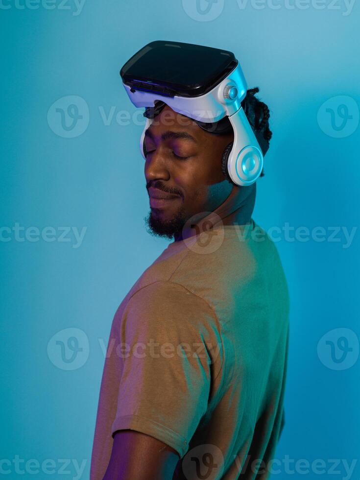 In a futuristic visual, an African American man stands isolated against a striking blue backdrop, adorned with VR glasses that transport him into a cutting-edge virtual reality experience, merging photo