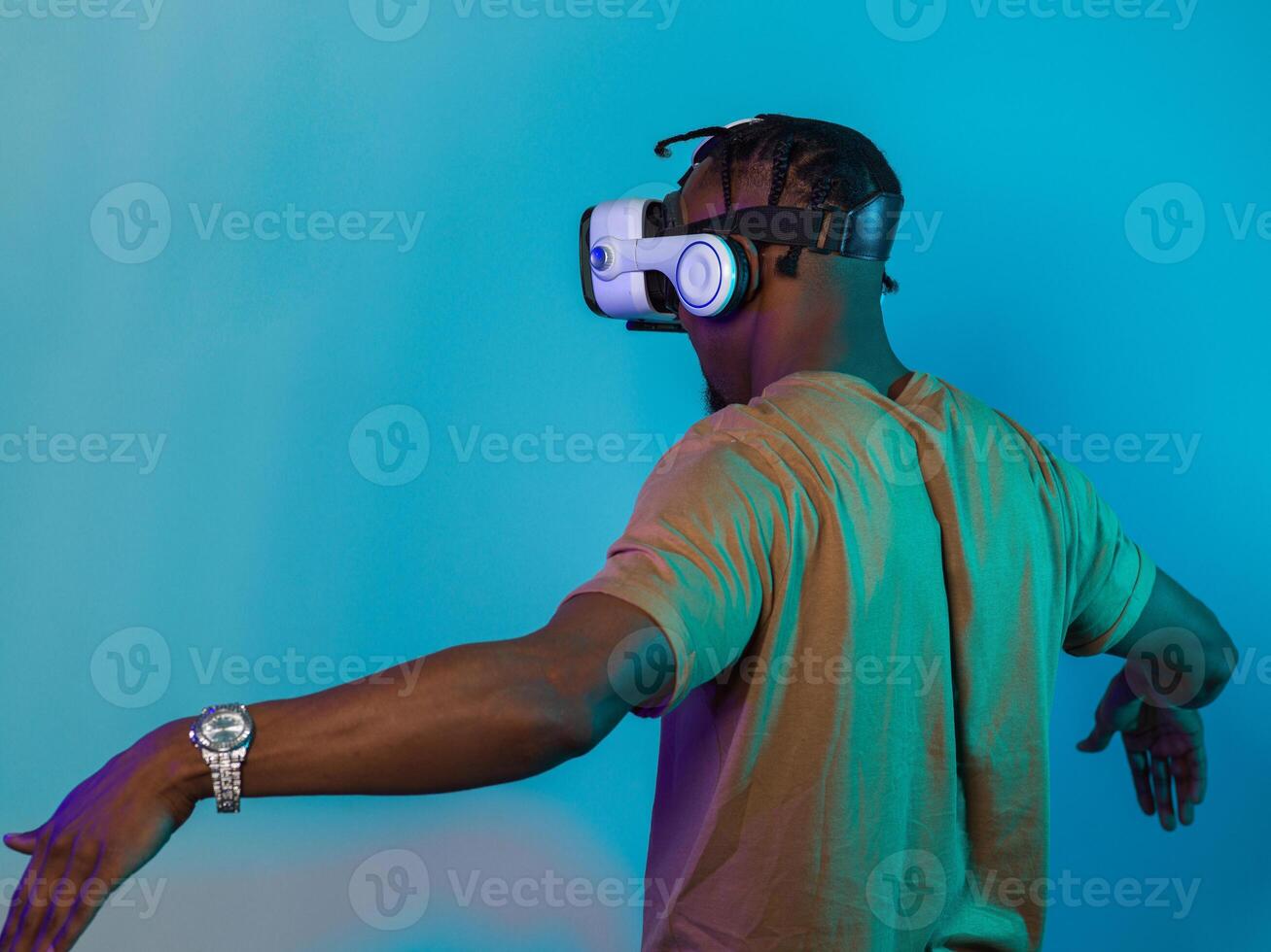 African American man dons VR glasses, engrossed in a virtual reality simulation, gesturing with hands, against a striking blue background, highlighting the fusion of cutting-edge technology and photo