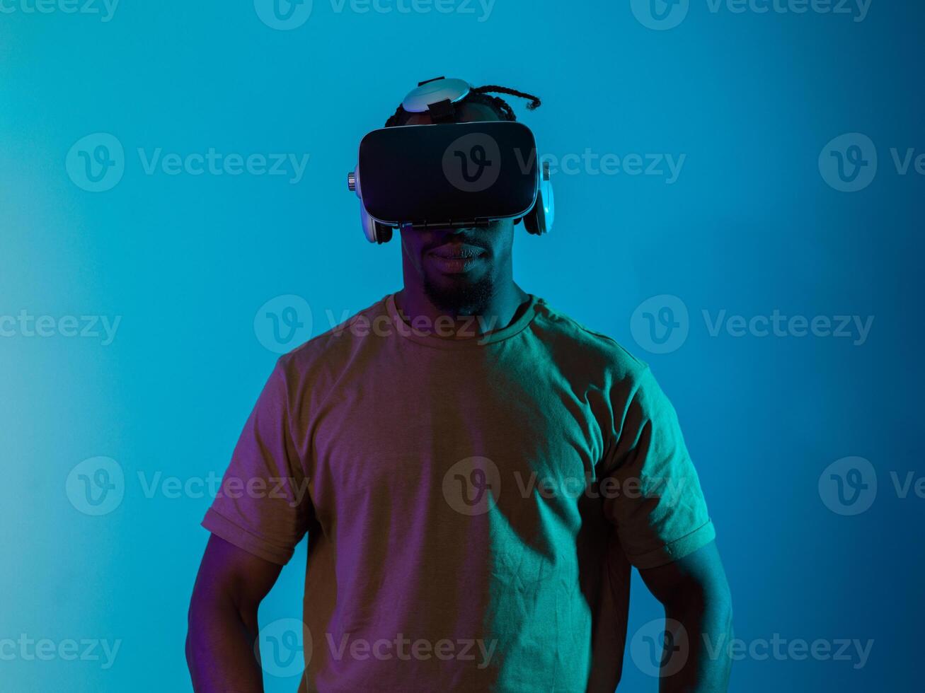 In a futuristic visual, an African American man stands isolated against a striking blue backdrop, adorned with VR glasses that transport him into a cutting-edge virtual reality experience, merging photo