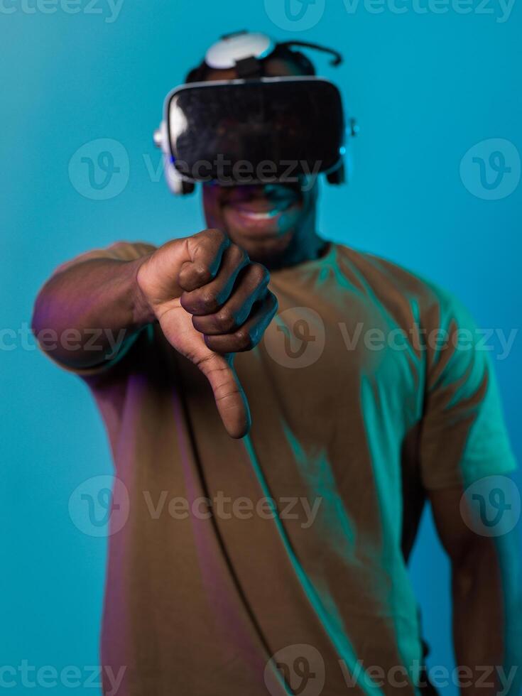 African American man, wearing virtual reality glasses, expresses dissatisfaction by pointing down with his hand while standing isolated against a blue background, portraying a modern critique within photo