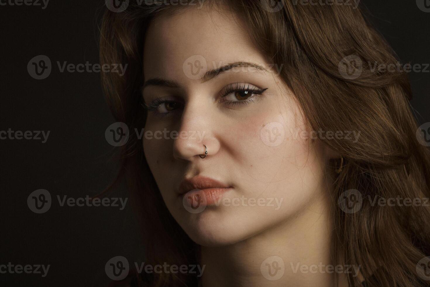 Portrait of a young brunette girl with a haughty look. Close-up, black background. piercing in the nose. photo