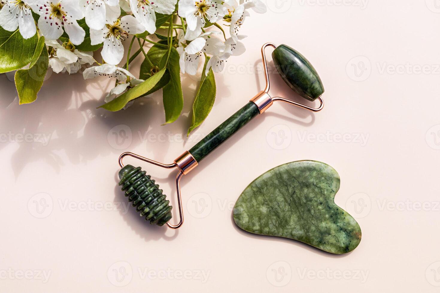 Jade Roller Massager and Gua Sha Scraper For Face And Neck Skin Massage On Beige Background With Flowering Fruit Tree Branch. home self-care. photo