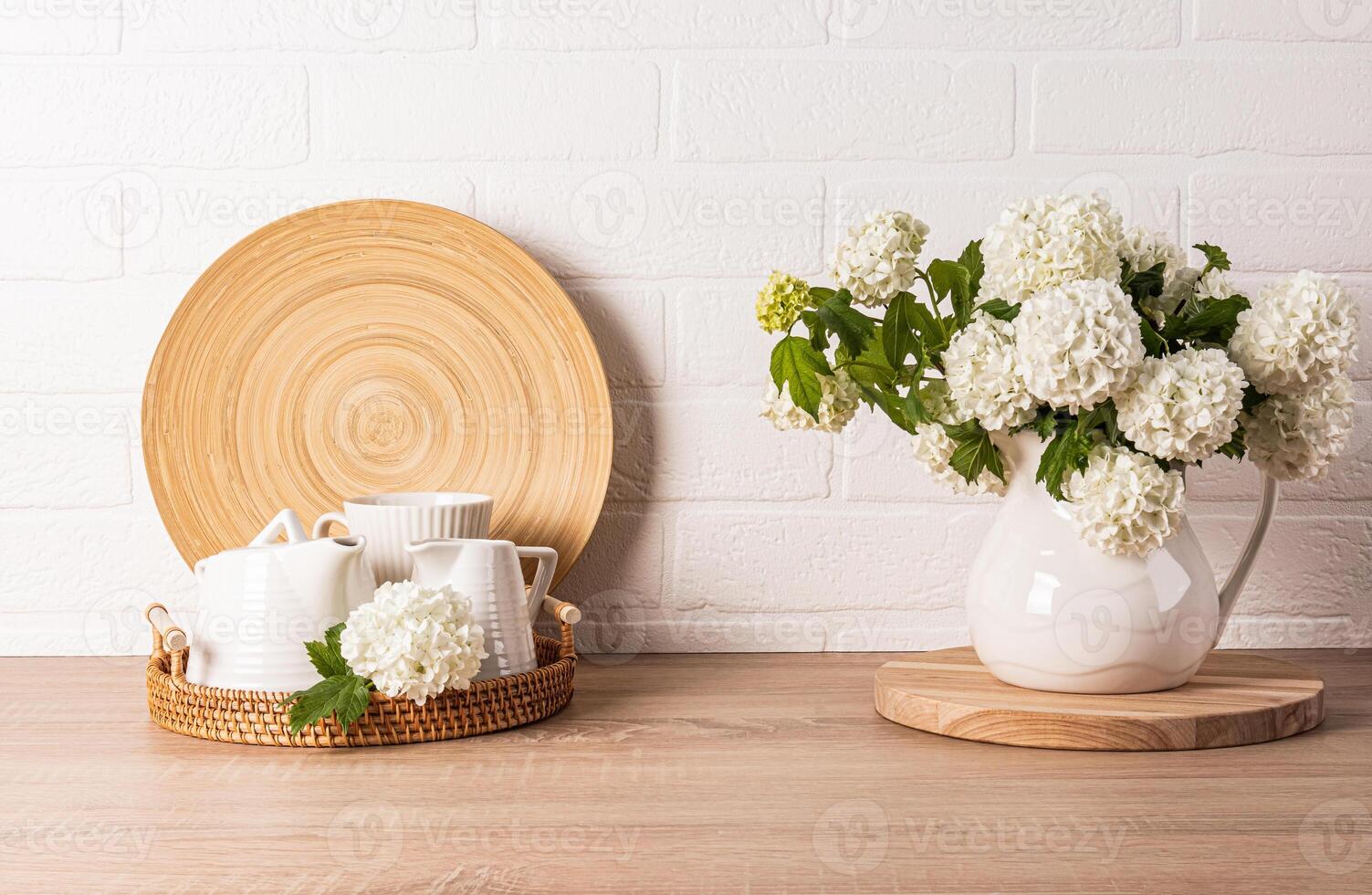White ceramic tea set on round wicker tray on kitchen countertop. decorative flowers in jug. Kitchen background in light colors. Front view photo