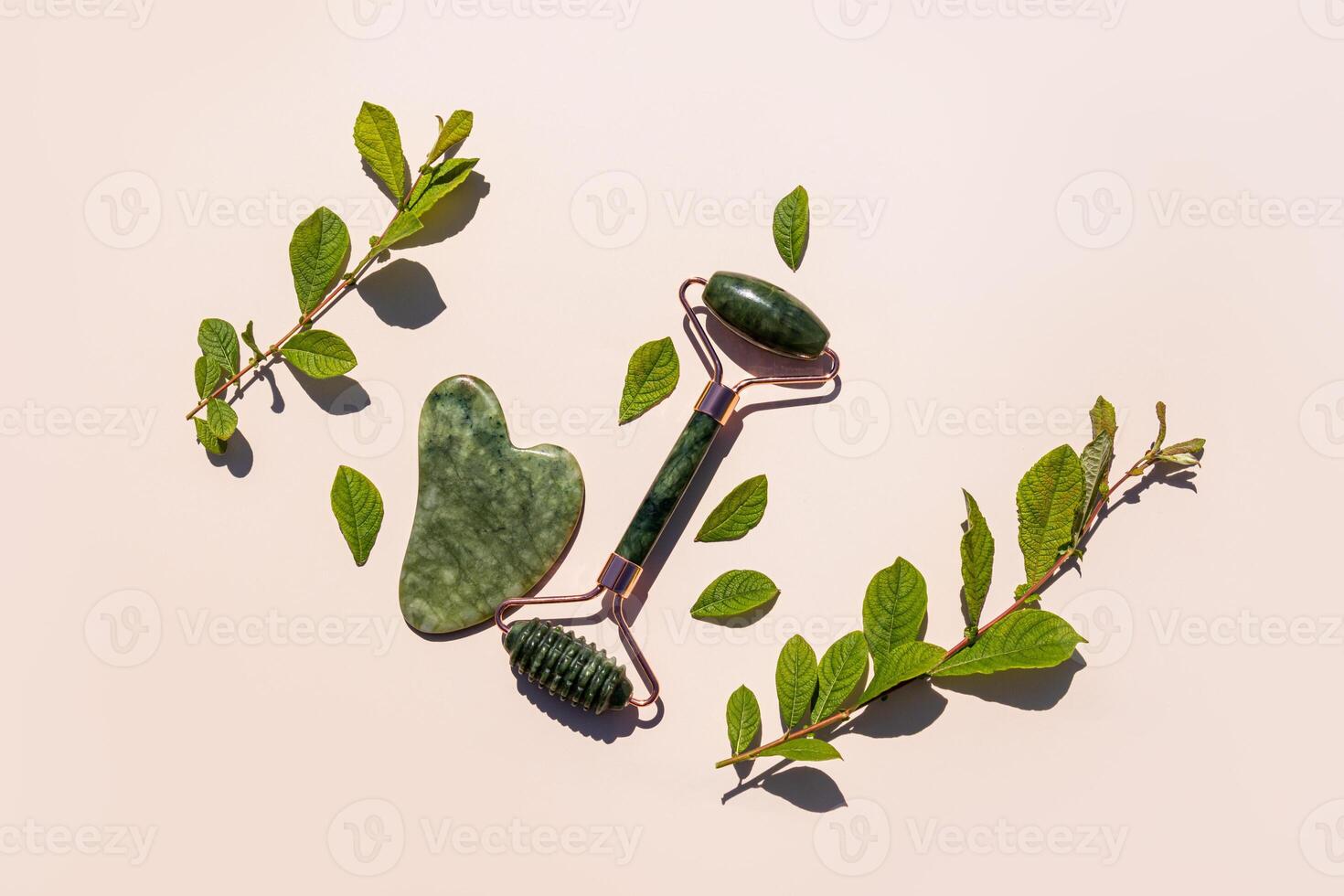 Green malachite roller massager and jade gua sha scraper on a beige background with green natural twigs. top view. flat lay. Natural care. photo