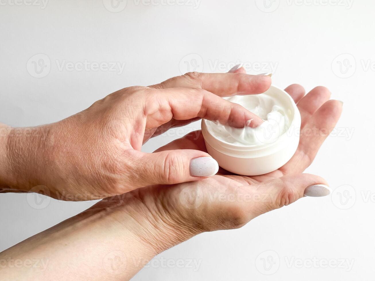Close up of middle age woman hands holding container of cream and dipping finger on white background. Skincare and moisturizing concept. For healthcare, wellness, and self care poster. photo