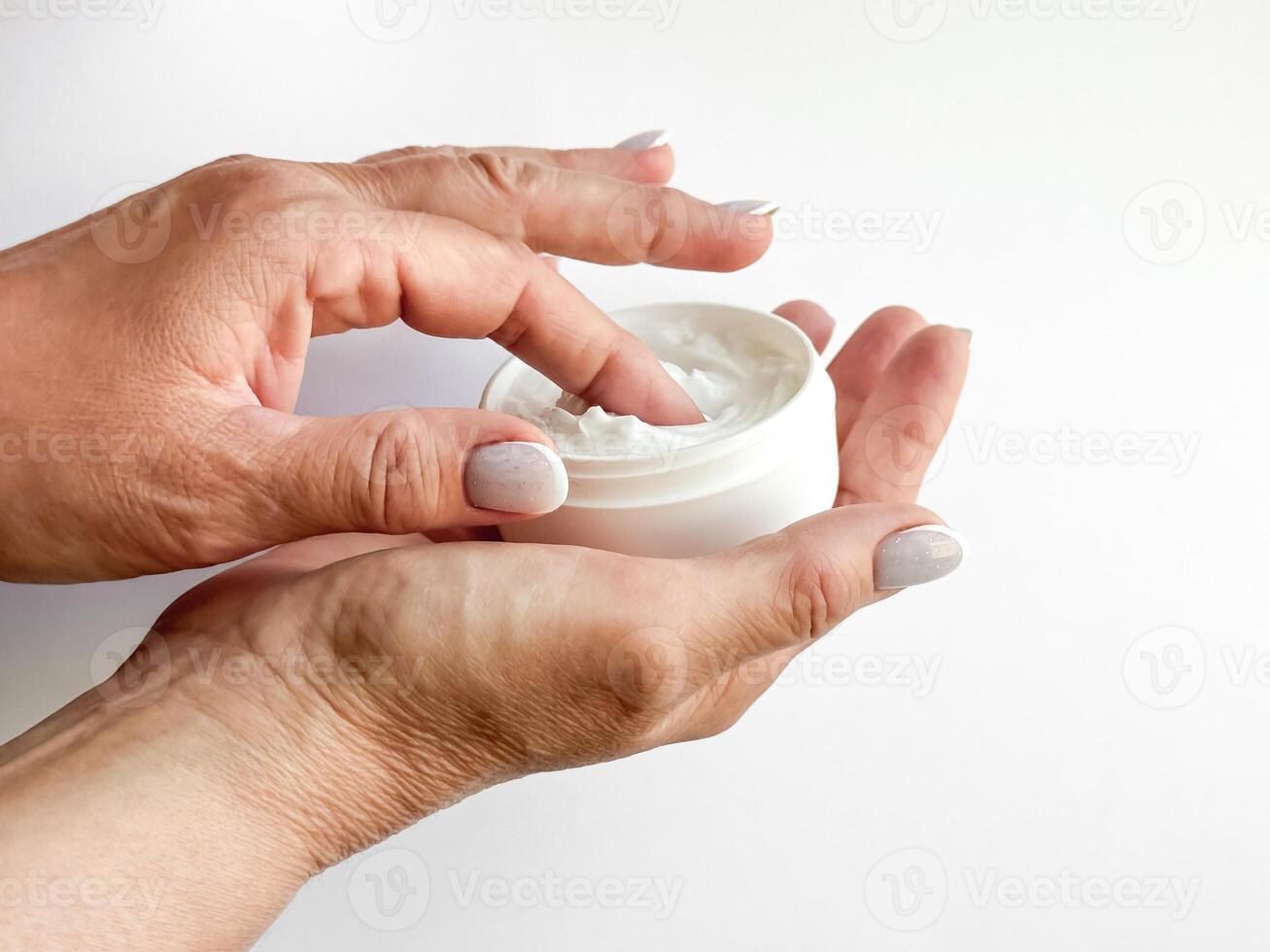 Close up of middle age woman hands holding container of cream and dipping finger on white background. Skincare and moisturizing concept. For healthcare, wellness, and self care poster. photo