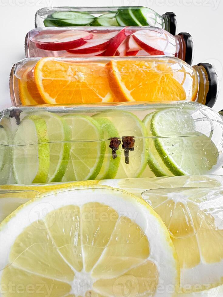 Stacked glass bottles with detox drinks with cucumber, apple, orange, and lemon slices on white background. Vertical orientation, refreshing beverage concept for design and print. photo