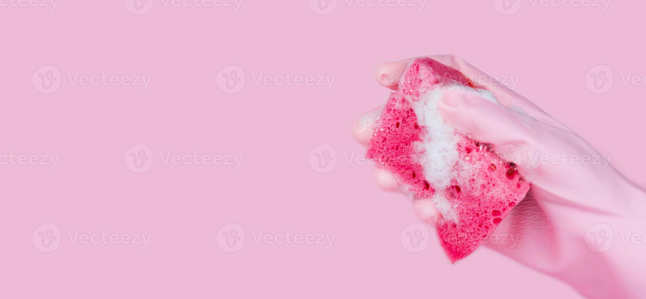A women's hand in pink rubber glove holds a cleaning sponge with soapy foam on a pink background. Copy space. Banner. Selective focus. photo