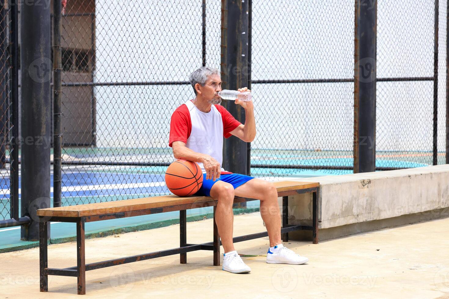 Senior Asian man in retirement age is drinking water after playing basketball at outdoor court for healthy and fun activity to exercise and workout against aging illness photo