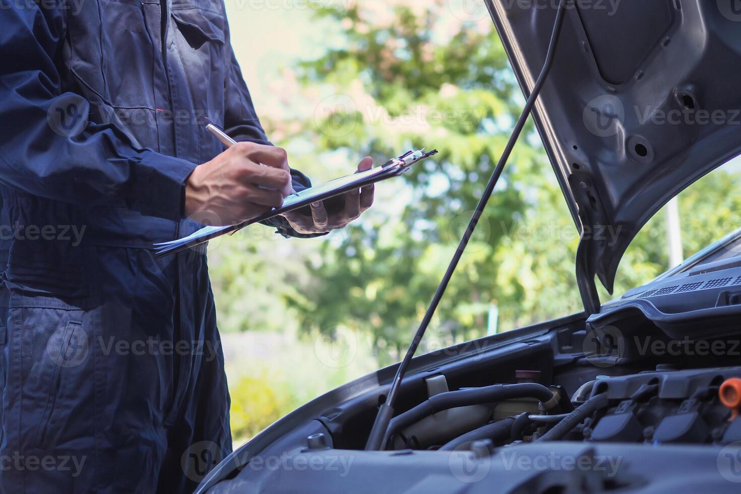 mechanic is writing on a clipboard next to a car. The car is parked in a parking lot with trees in the background photo