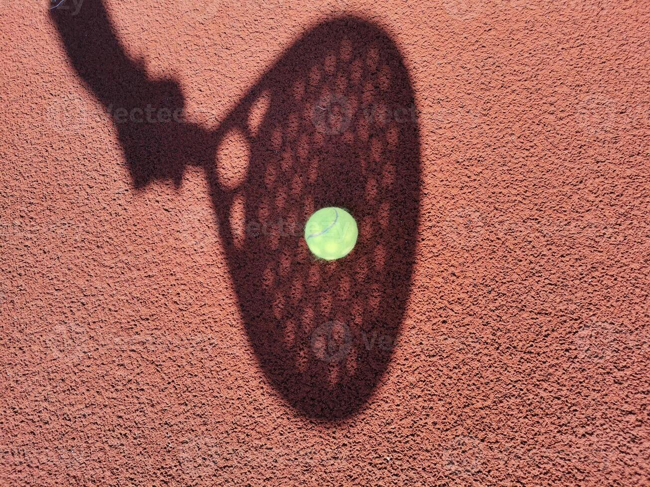 High angle of yellow ball placed on court and covered with shadow of crop person holding padel racket photo