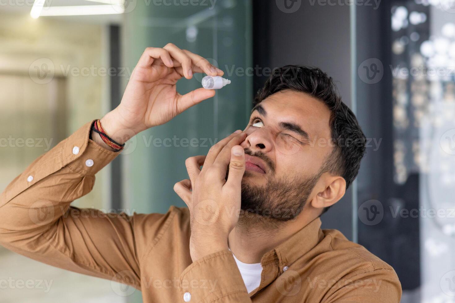 Close-up photo of a young Indian man standing in a modern office and instilling medicine for fatigue and inflammation into his eye.