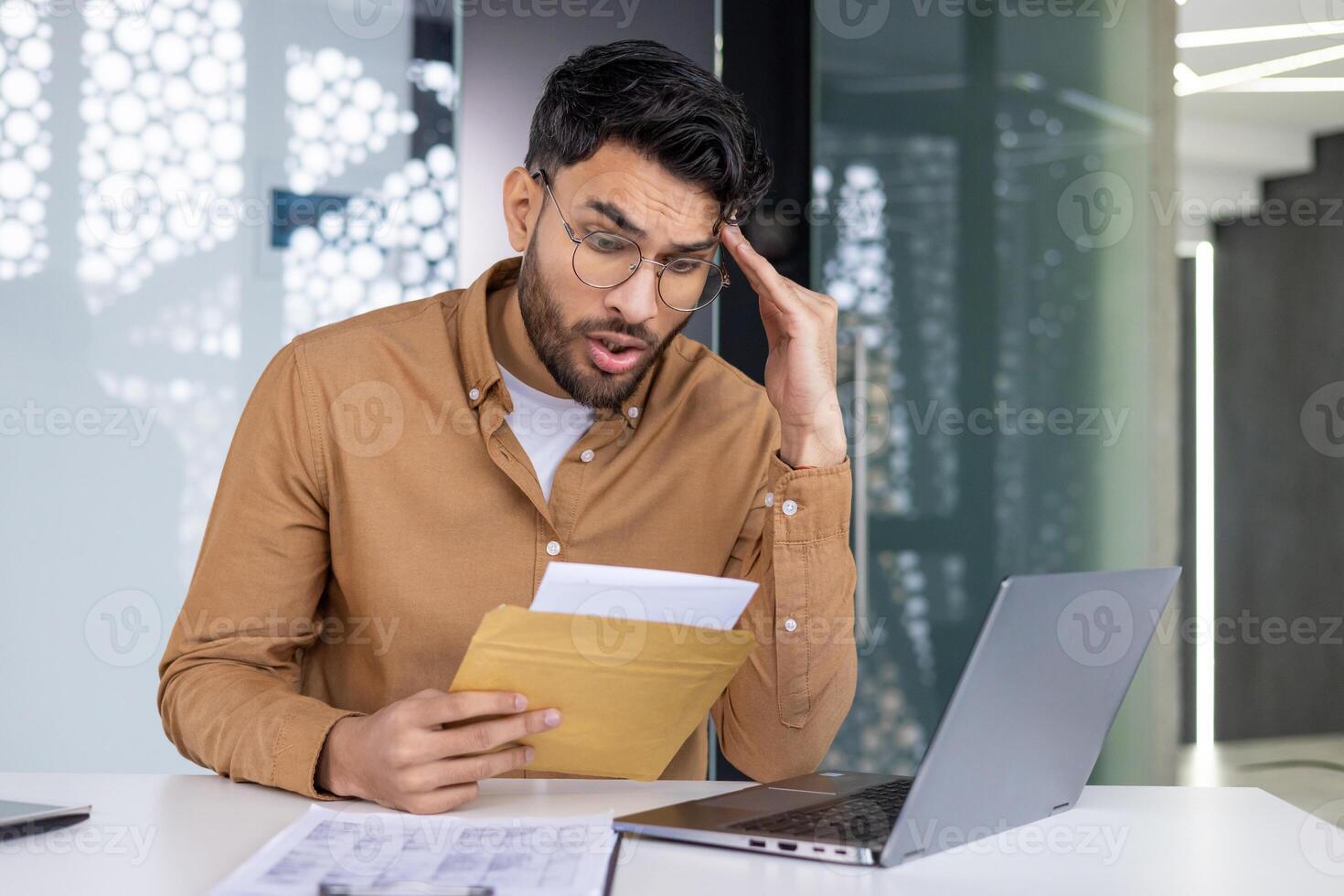Shocked man reads a letter with bad news at the office while working on his laptop. Business concern and stress. photo