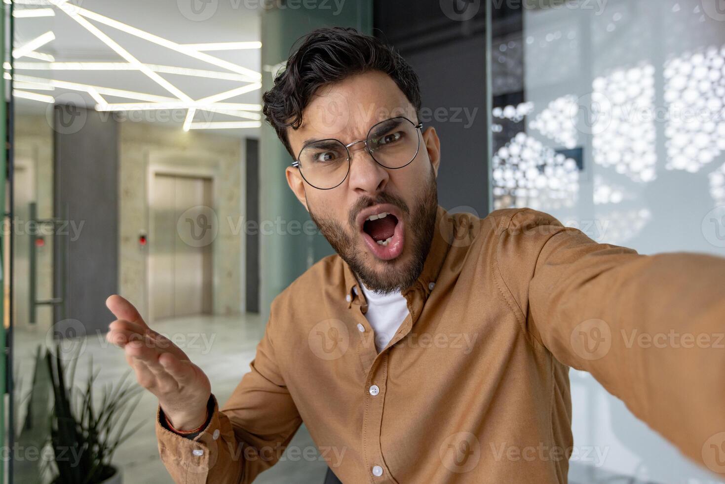 Close-up photo of an angry young Indian man in glasses who is in the office and emotionally talking on a call to the camera.