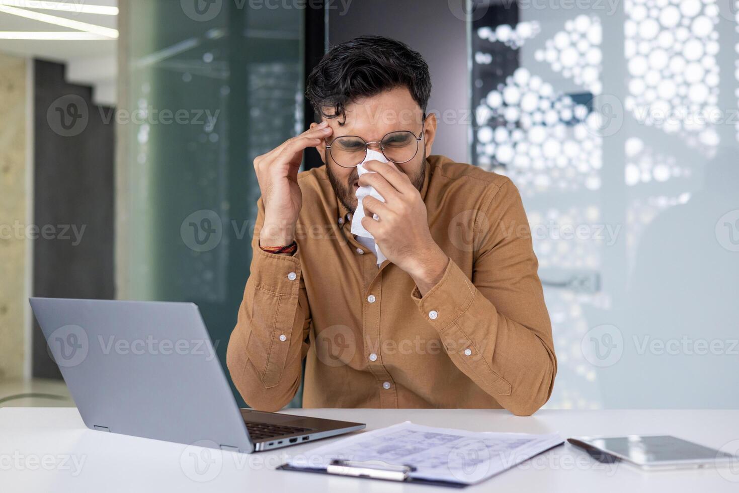 Man struggling with cold symptoms while trying to work on his laptop in the office. Concept of illness, health issues, and work environment. photo