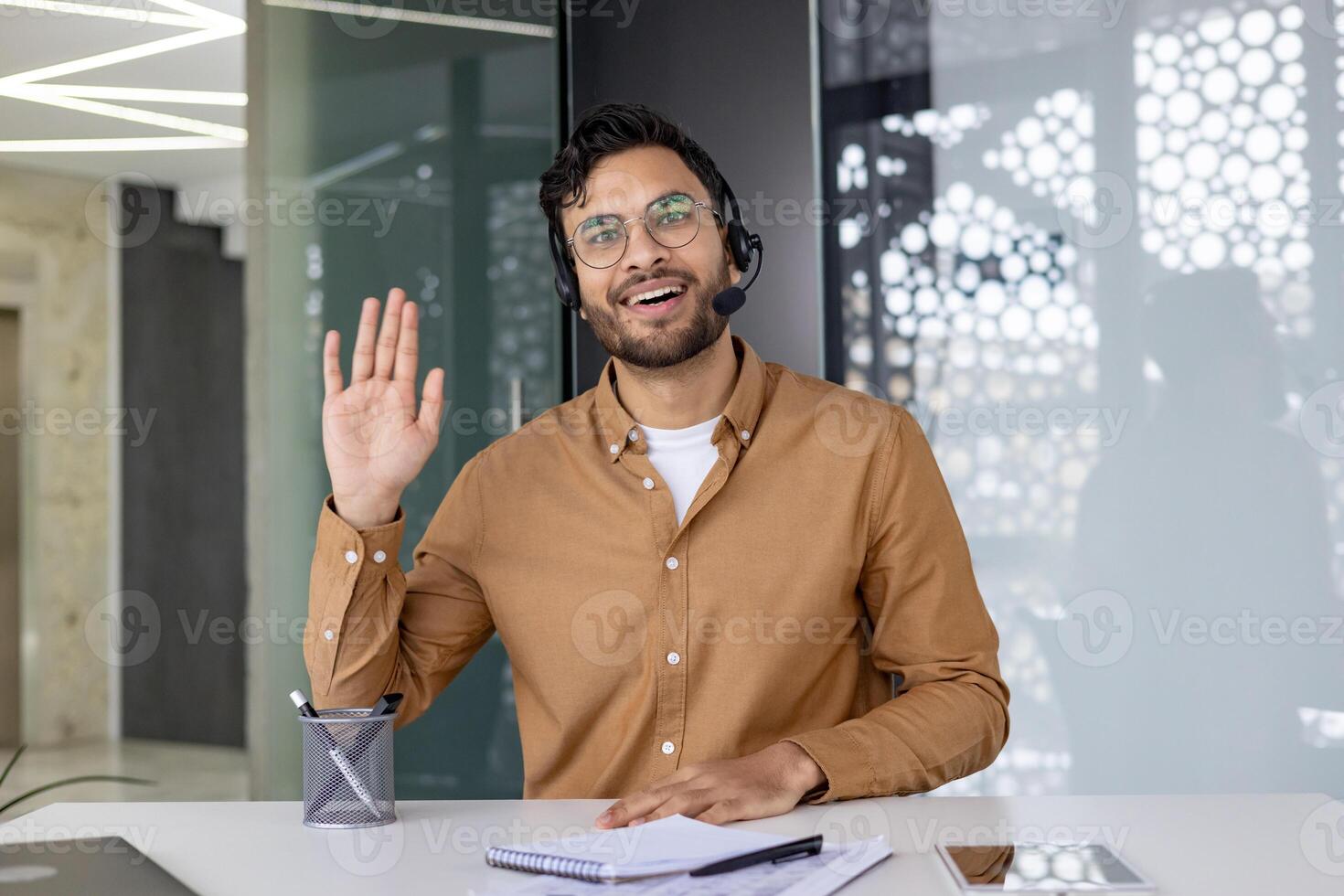Happy man wearing a headset and waving while looking at the camera during a chat in a modern office setting. photo