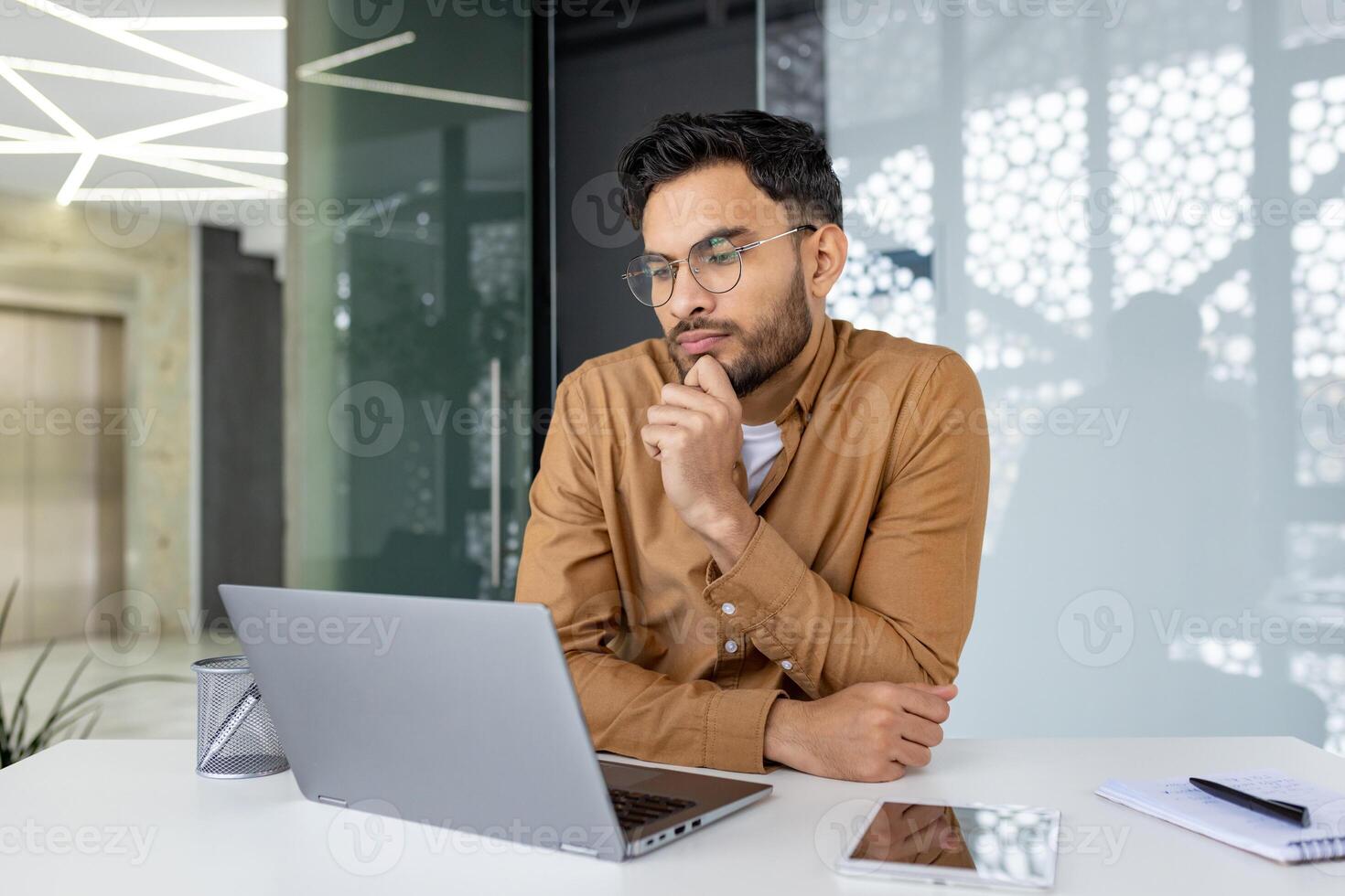 Young professional with glasses working on a laptop at a desk in a modern office. Considerate expression, focused on computer screen. photo