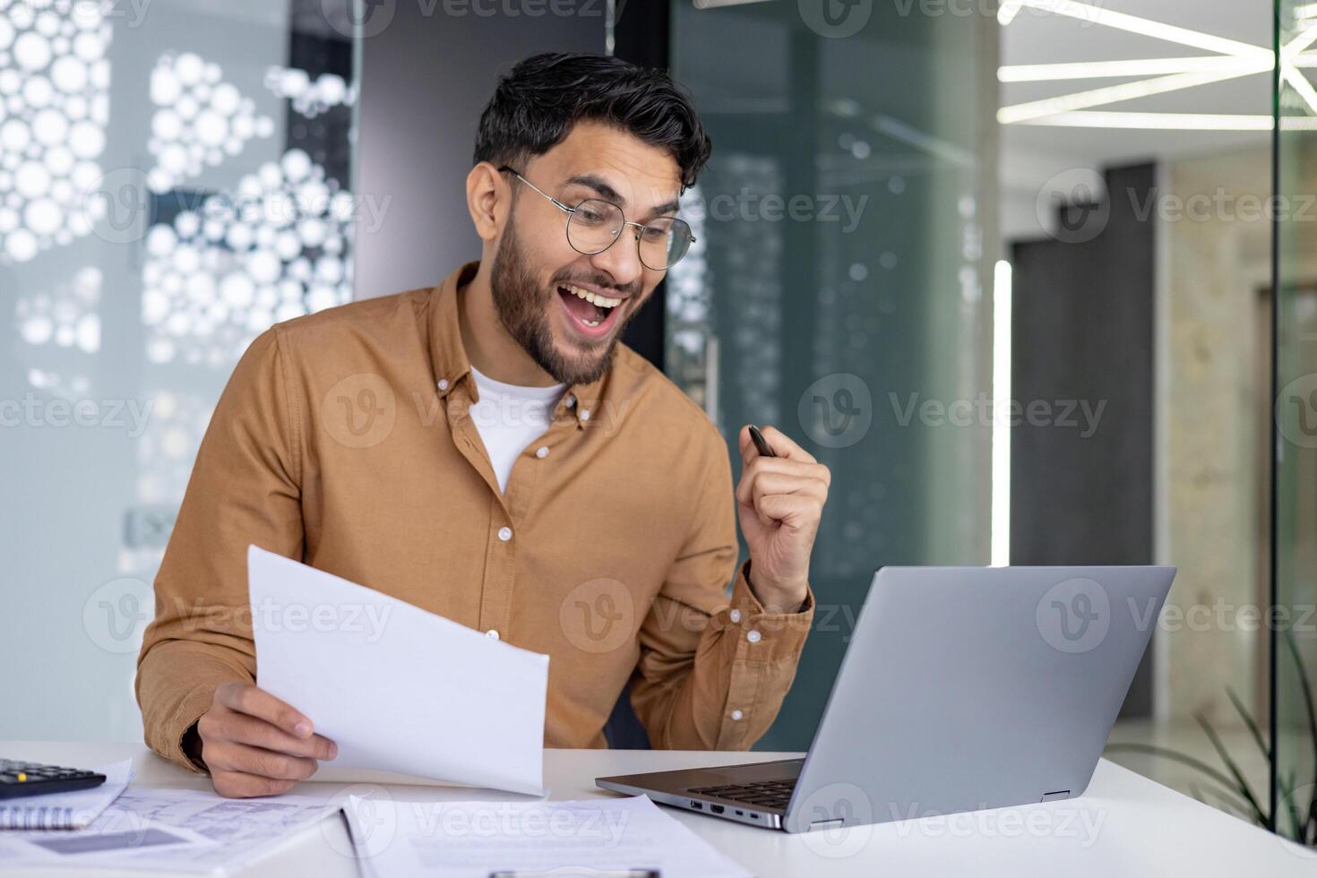 Happy Indian man sitting in office at desk, holding documents in hand, looking at laptop screen and happy with achievement and result. photo
