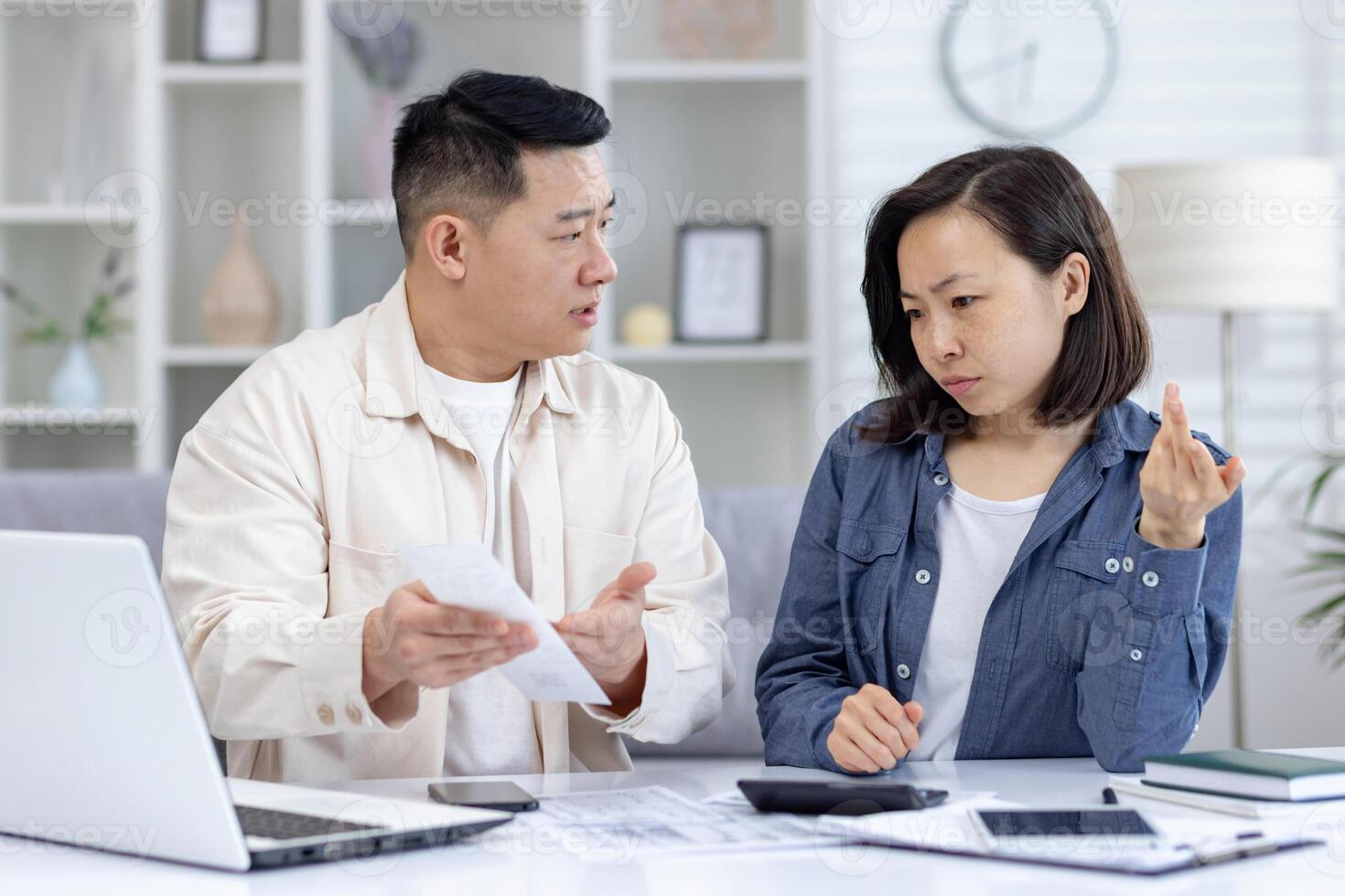 Couple having a serious discussion about finances at home, with bills and a laptop on the table. Concern and tension are visible. photo