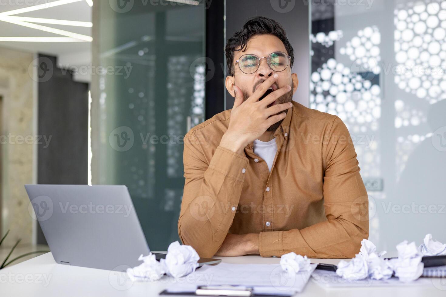 Tired businessman yawning at his desk surrounded by a laptop and crumpled papers in a modern office setting. Concept of fatigue, overworked, and stress. photo