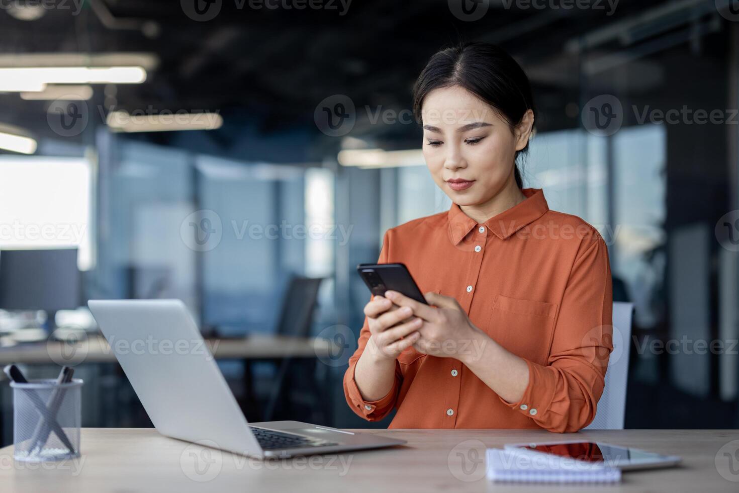 Asian businesswoman using smartphone at office desk, working with laptop, and writing in a notebook in a modern office space. photo