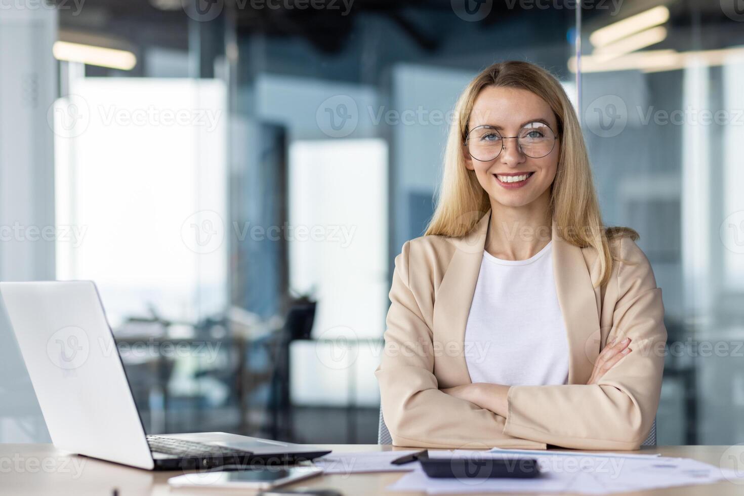 Portrait of a successful and self-confident business woman in a business suit, sitting in the office at the desk and looking at the camera, smiling with her arms crossed. photo