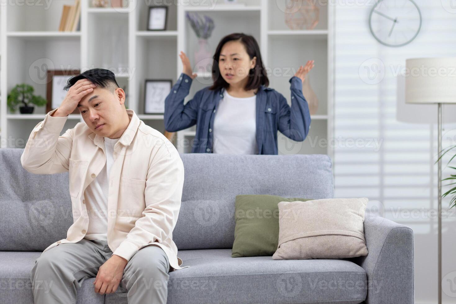 A stressed couple arguing at home in a modern living room. The man is sitting on the couch, while the woman stands behind with raised arms. photo