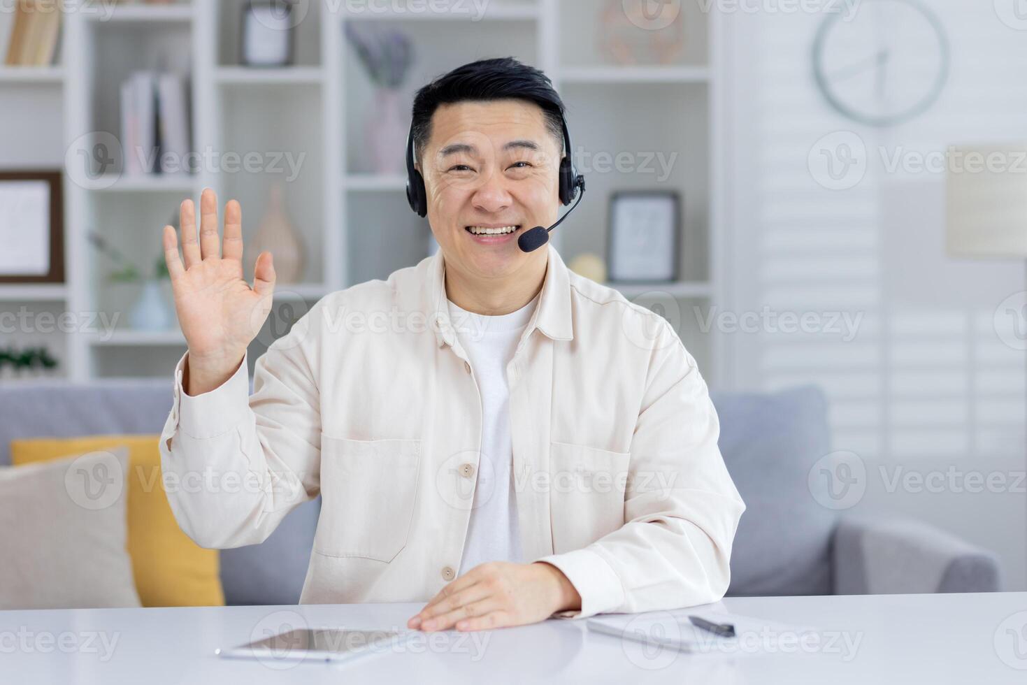 A cheerful Asian man wearing a headset smiles and waves at the camera, sitting comfortably at his home office with decor in the background. photo
