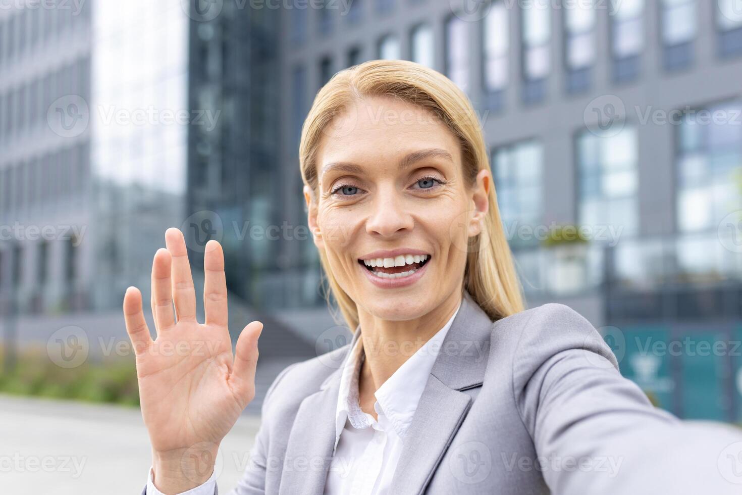 A cheerful businesswoman in a suit waving and smiling at the camera in front of a contemporary office building. photo