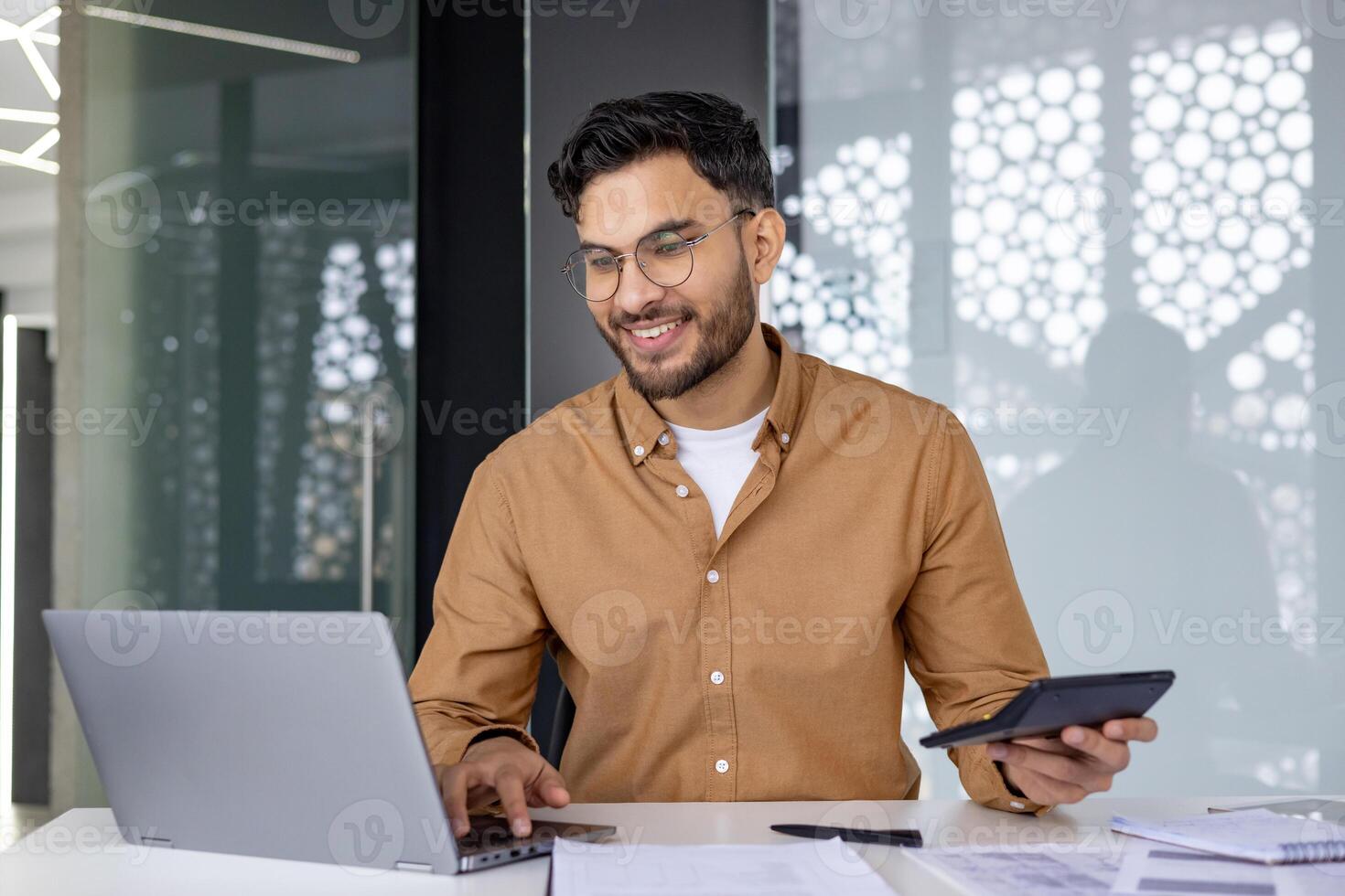 Financier working on auditing paperwork, using a calculator and laptop in a modern office setting, showcasing a productive work environment. photo