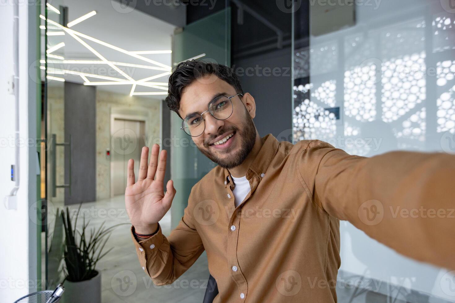 Smiling man wearing glasses and waving at camera in a modern office setting during a call. Friendly and approachable atmosphere. photo