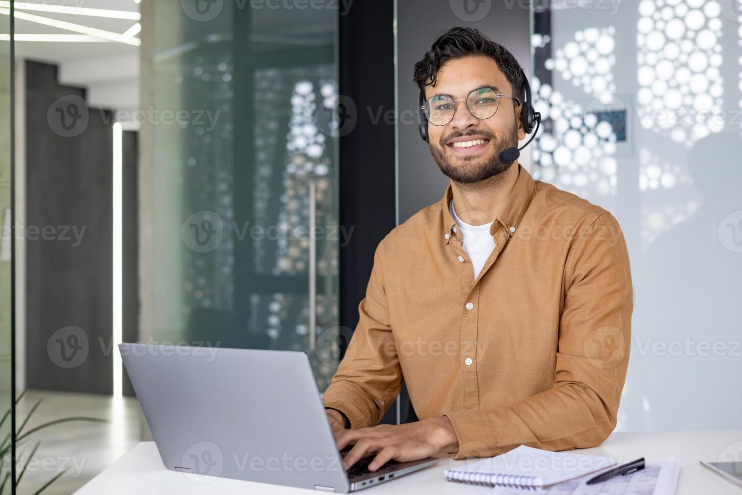 Portrait of a smiling young Muslim man wearing a headset, working in the office at a table on a laptop, looking confidently at the camera. photo